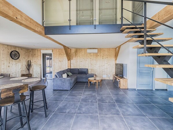 A modern living space with a wooden interior, a gray sofa, a dining area with a round table and chairs, and a staircase with a glass railing.