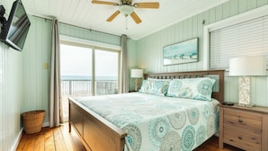 Ein Schlafzimmer mit einem Holzbett, einem Deckenventilator, einem Nachttisch mit Lampe und einem Fenster mit Blick aufs Meer.