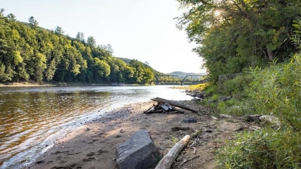 A riverbank with a wooden bridge, dense forest, and a rocky shoreline. - Cozy Adirondack Escape (Warrensburg)