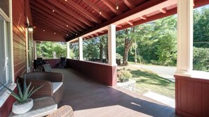 A covered porch with wicker furniture, a potted plant, and a view of a green landscape. - Cozy Adirondack Escape (Warrensburg)