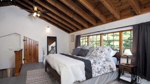 A bedroom with a large bed, wooden ceiling beams, a window with curtains, and a nightstand with a lamp. - Cozy Adirondack Escape (Warrensburg)