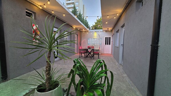 A narrow outdoor corridor with a table and chairs, string lights, and potted plants.