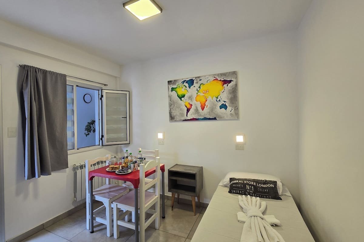 Une petite pièce bien éclairée, avec une table à manger, des chaises, un petit meuble et un lit avec une serviette.