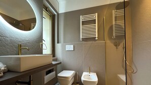 Une salle de bain moderne avec un grand miroir ovale, un lavabo sur colonne, des toilettes, un bidet et une douche à l’italienne.
