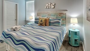 A bedroom with a bed, a wooden headboard, a wall clock, a small round table with a lamp, and a door. - Port A's Flip Flops & Pop Tops by Silver Sands Vacation Rentals (Port Aransas)