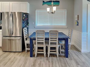 Une cuisine équipée d’un réfrigérateur en acier inoxydable, d’une table à manger en bois avec des chaises, d’une fenêtre avec des stores et d’une décoration murale représentant des poissons.