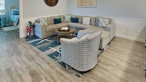 A living room with a sectional sofa, a round coffee table, a patterned rug, a wall clock, and a map. - Port A's Flip Flops & Pop Tops by Silver Sands Vacation Rentals (Port Aransas)