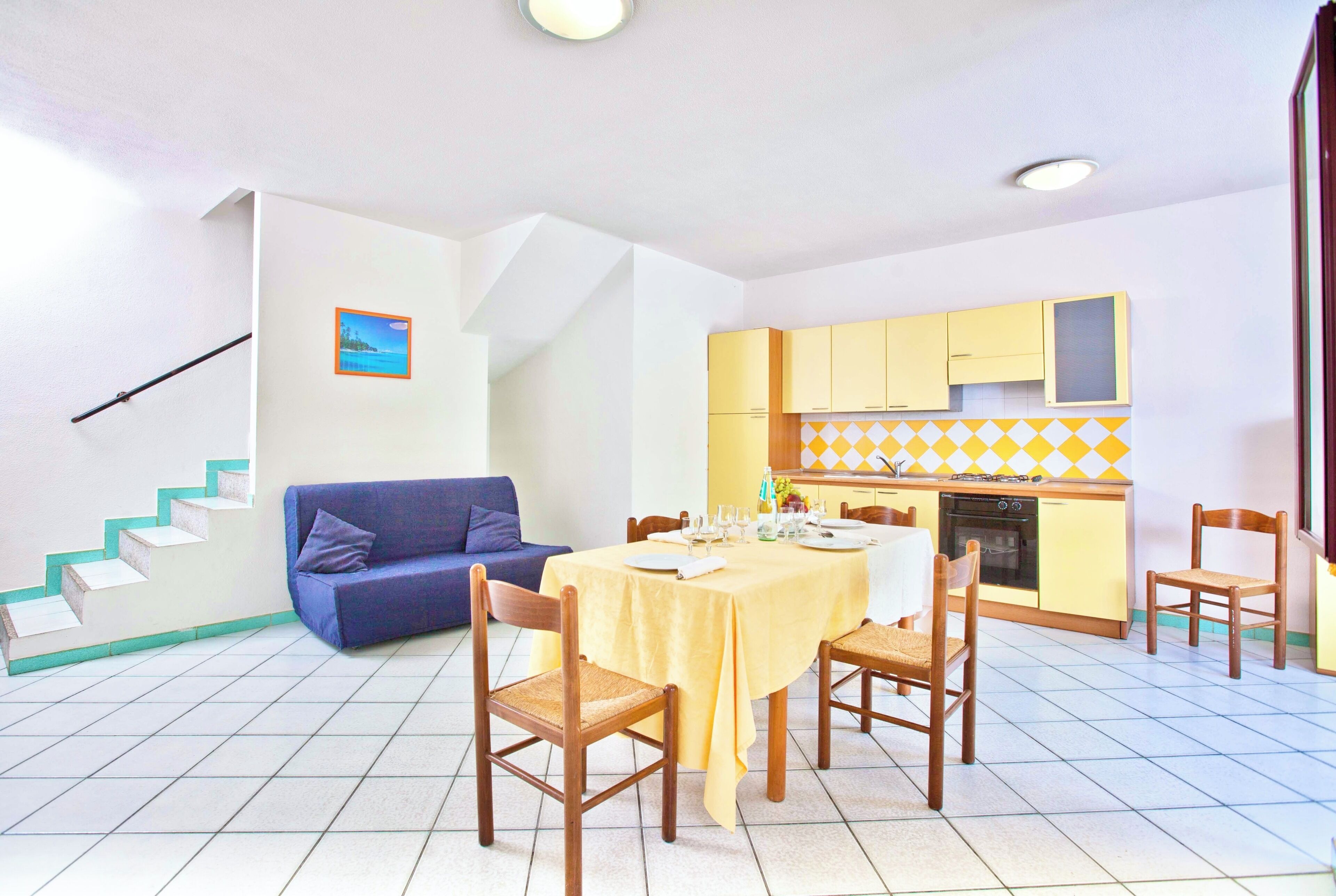 A dining area with a table set for four, a blue sofa, wooden chairs, and a kitchen in the background.