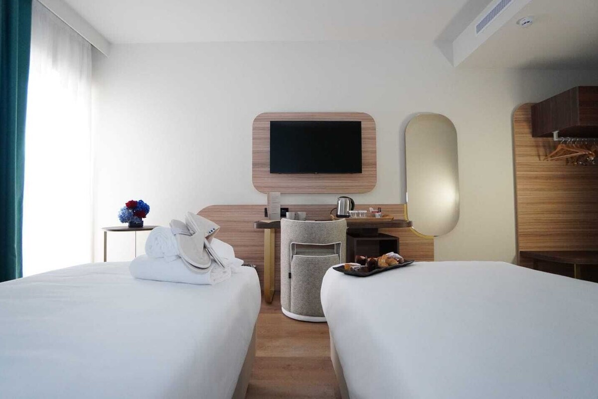 A hotel room with two beds, a TV, a desk with a chair, and a small table with a vase of flowers.