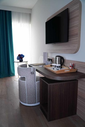 Ein kompaktes Hotelzimmer mit einem an der Wand befestigten Flachbildfernseher, einem kleinen Tisch mit Tablett, auf dem Gläser und ein Wasserkocher stehen, einem grauen Sessel und einem Mülleimer.