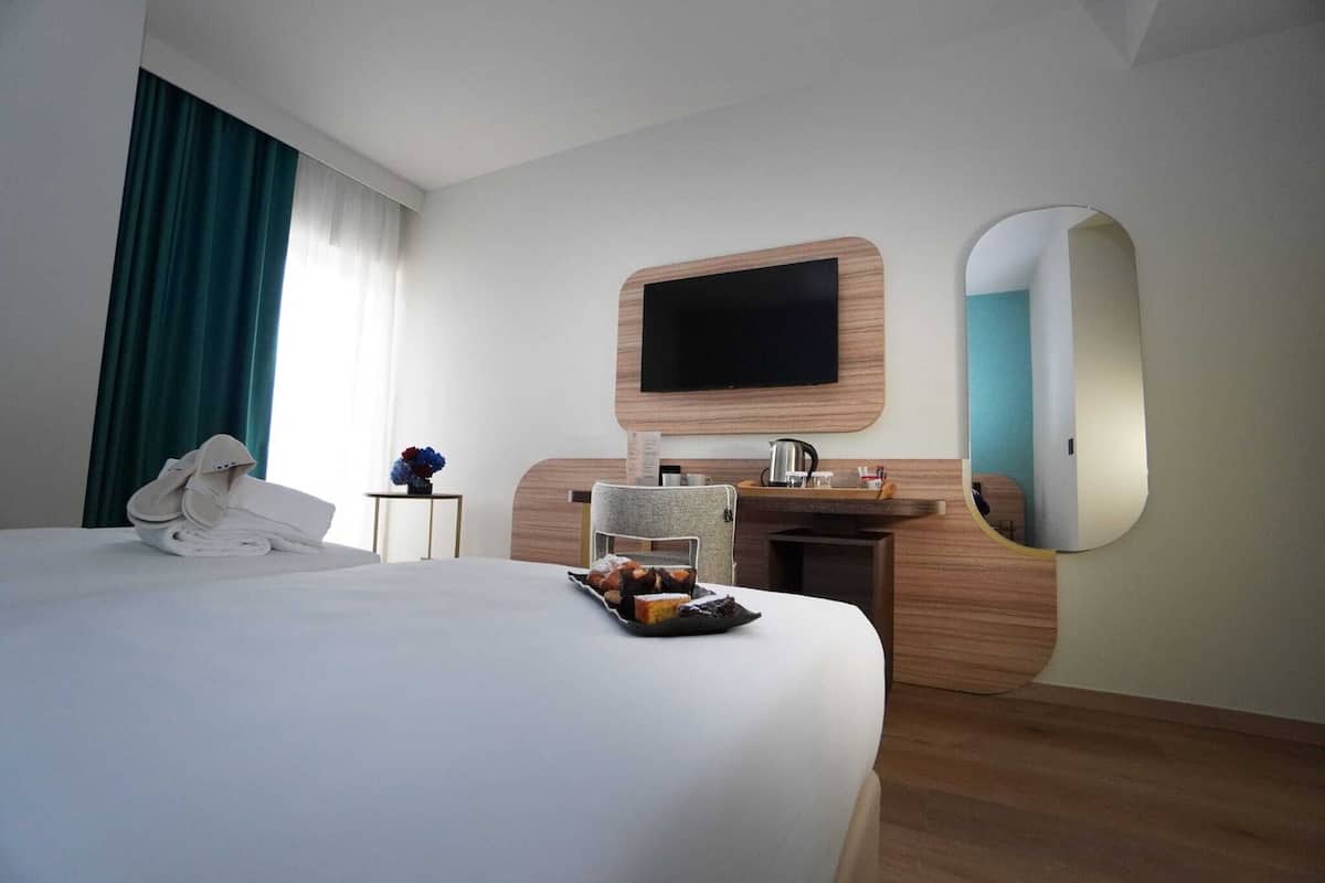 Ein Hotelzimmer mit einem großen Bett, einem flachbildfernseher an der Wand, einem kleinen Schreibtisch mit Spüle und einem Spiegel.