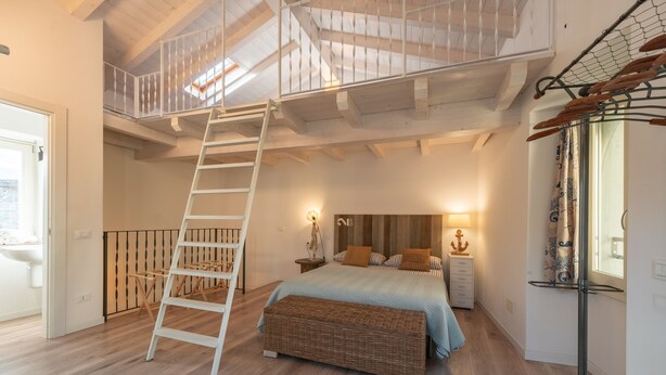 A bedroom with a wooden ladder leading to a loft, a bed with a blue bedspread, and a wicker bench. - Casa di Tully in Sarnico (Sarnico)