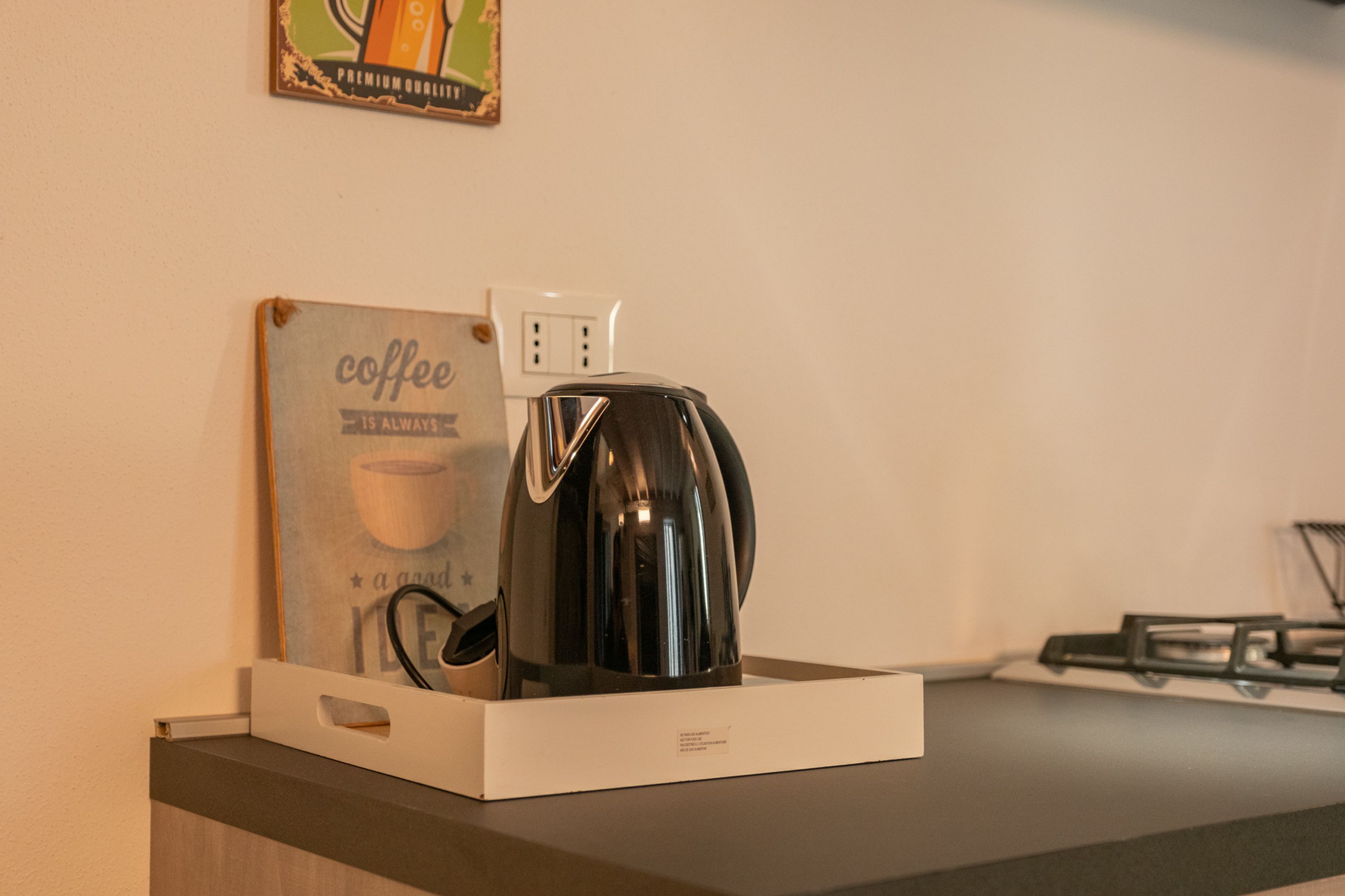 Una cafetera sobre una encimera, con un cartel con motivos de café y una toma de corriente al fondo.