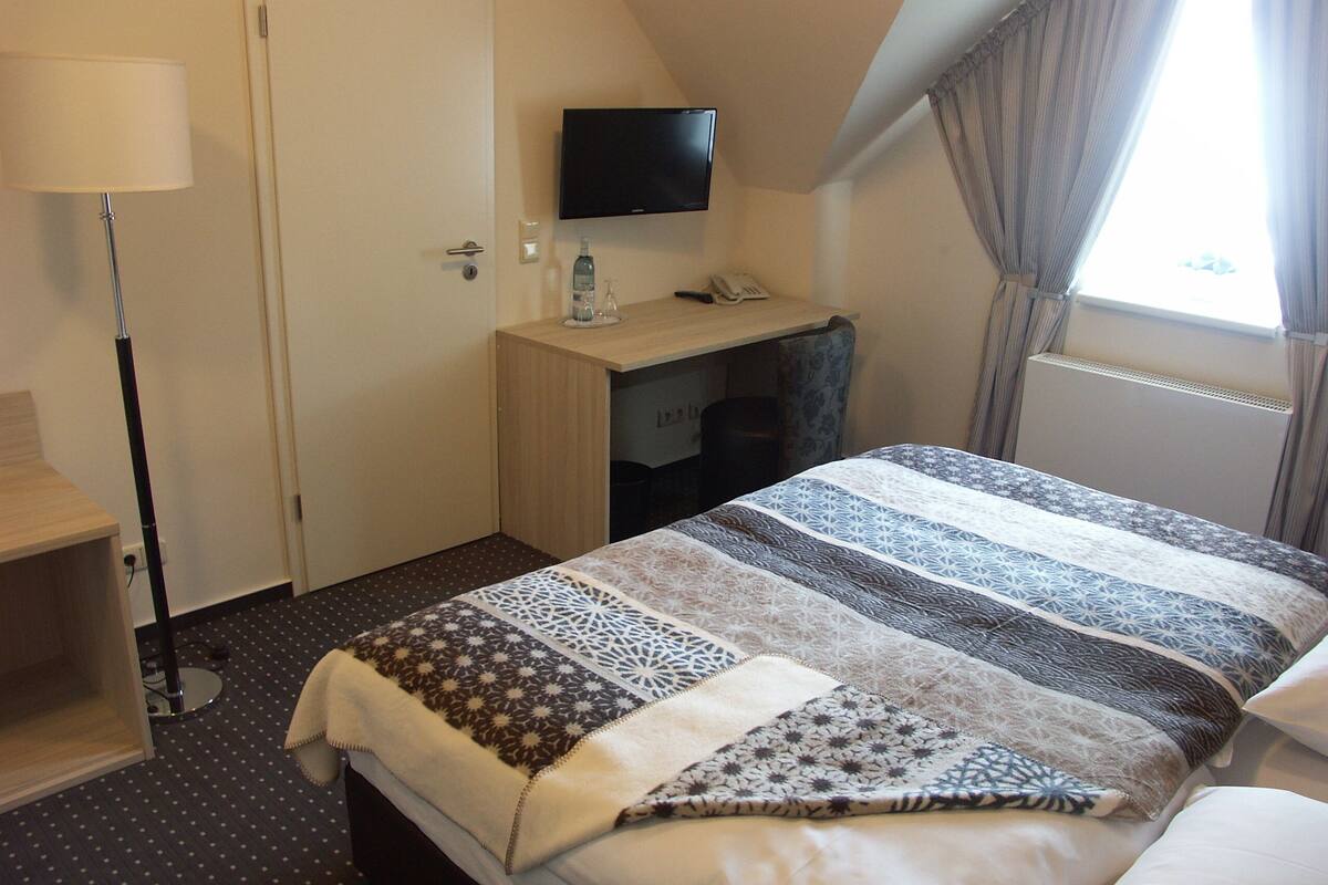Una habitación de hotel con cama, escritorio, televisor y ventana con cortinas.