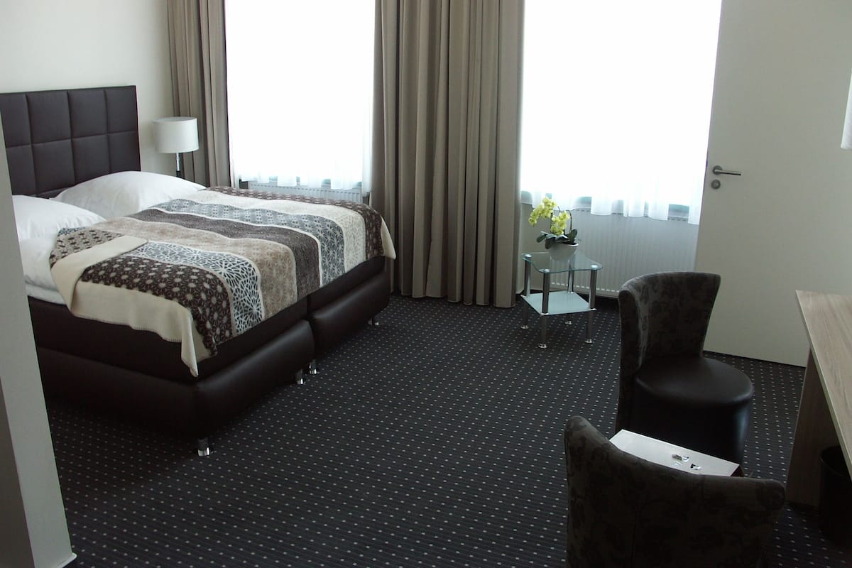 Una habitación de hotel con una cama grande, una mesita con un florero, una silla y una puerta.