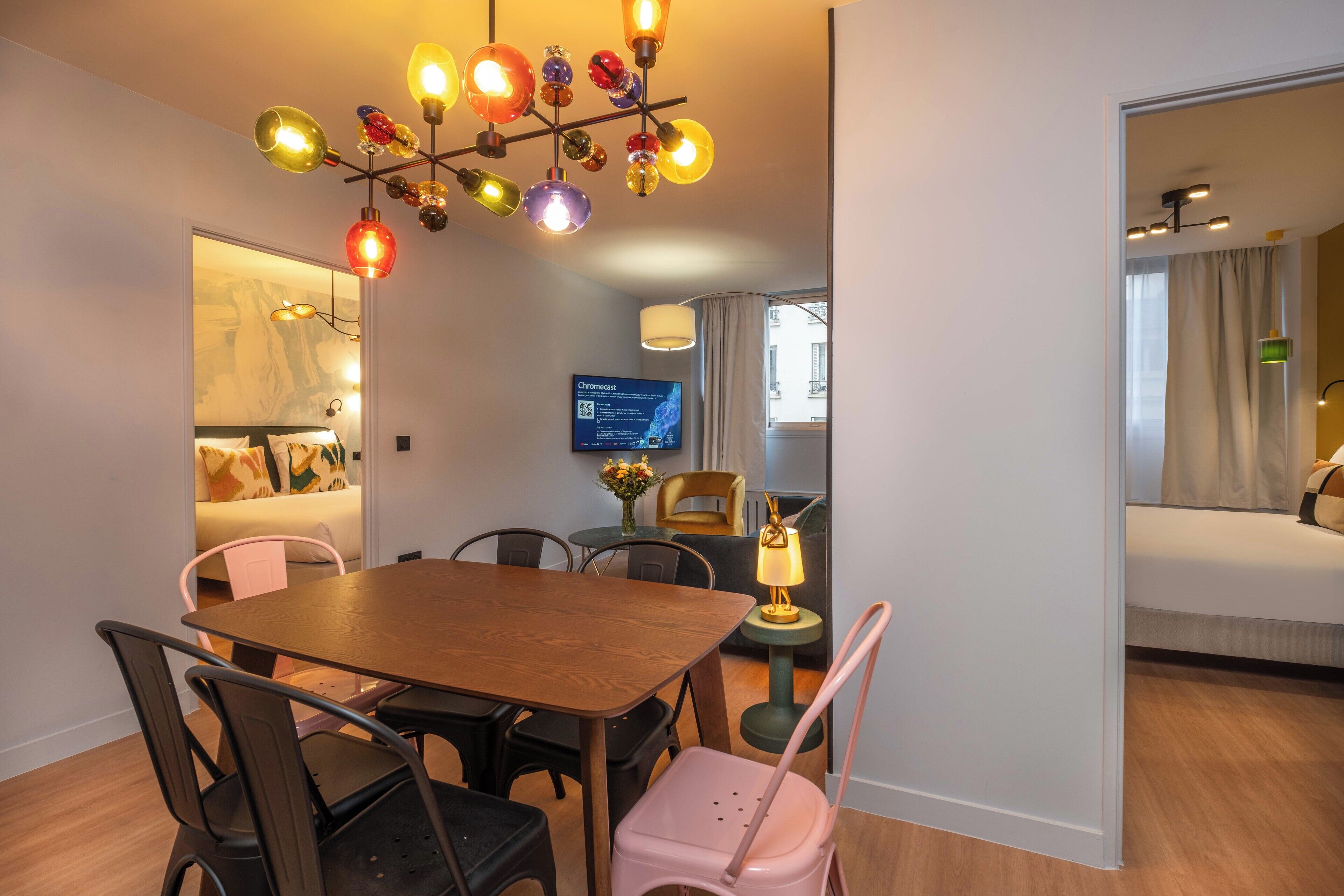 Habitación de hotel moderna con una mesa de comedor de madera, sillas negras y rosa, una lámpara de araña colorida, un televisor de pantalla plana y una cama con vista a la ventana.