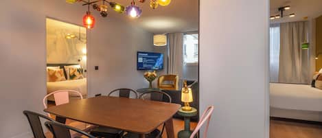 Habitación de hotel moderna con una mesa de comedor de madera, sillas negras y rosa, una lámpara de araña colorida, un televisor de pantalla plana y una cama con vista a la ventana.