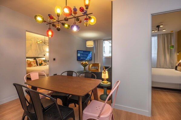 A modern hotel room with a wooden dining table, black and pink chairs, a colorful chandelier, a flat-screen TV, and a bed with a window view.