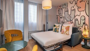 Habitación de hotel moderna con una cama, sillas amarillas, una mesita y una pared decorada con arte abstracto.