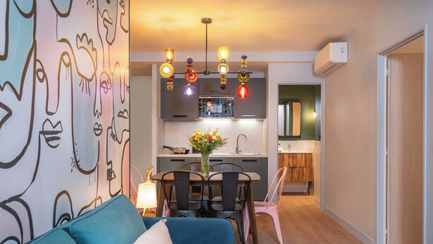Una sala de estar moderna con un sofá azul, una mesa de comedor con sillas, una zona de cocina con un fregadero y armarios, y una llamativa obra de arte en la pared.
