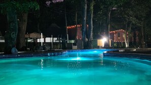 A swimming pool with a tiled edge, surrounded by trees and a building with string lights.