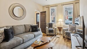 A living room with a grey sofa, patterned armchairs, a coffee table, and a TV stand. - Stylish Family Retreat | Near Downtown & Street Parking (Pittsburgh)