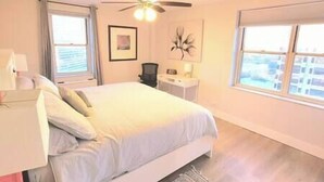 A bedroom with a bed, a desk, a chair, a ceiling fan, and two windows.