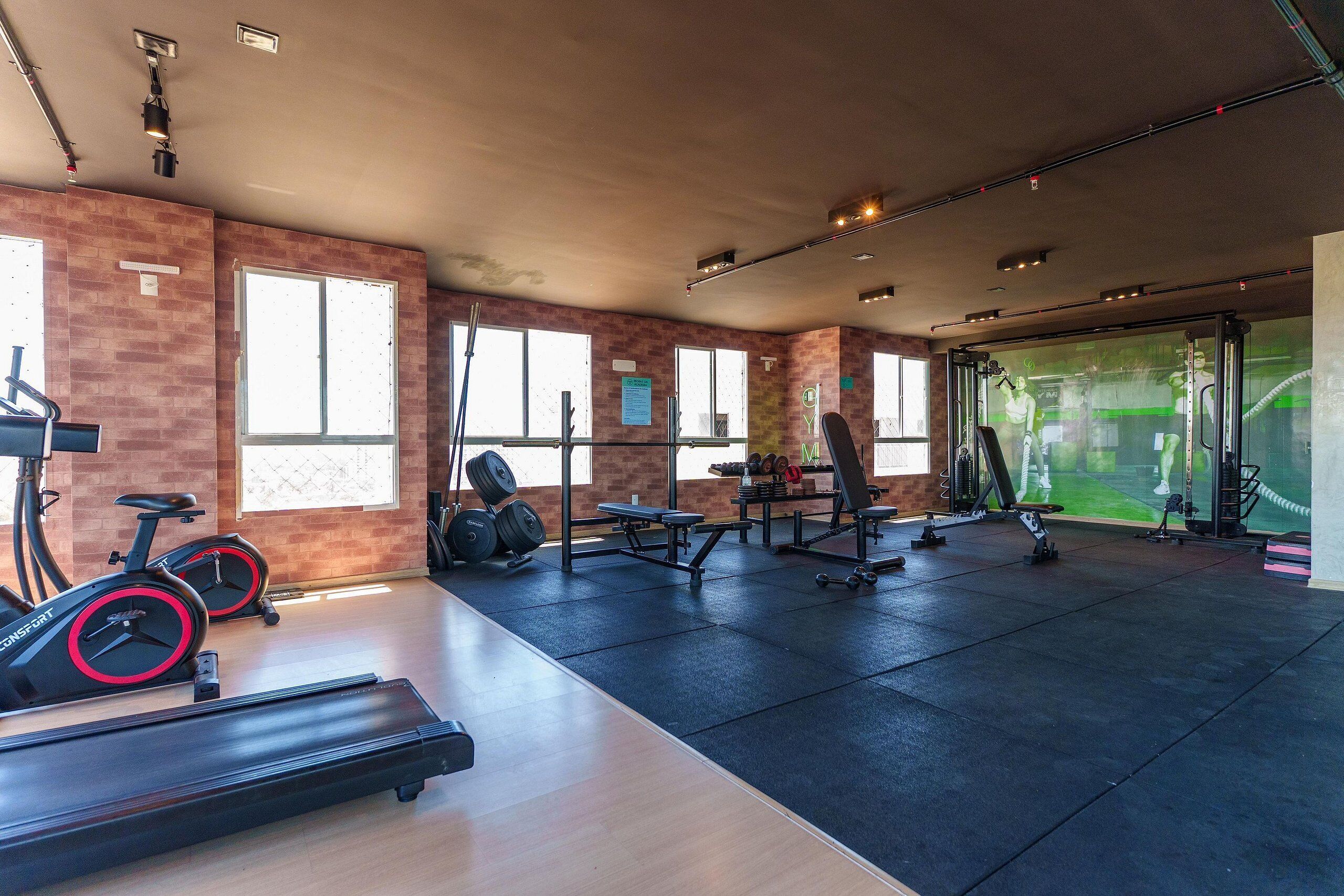 Un gimnasio con equipo de ejercicio, ventanas amplias y una pared de ladrillo.