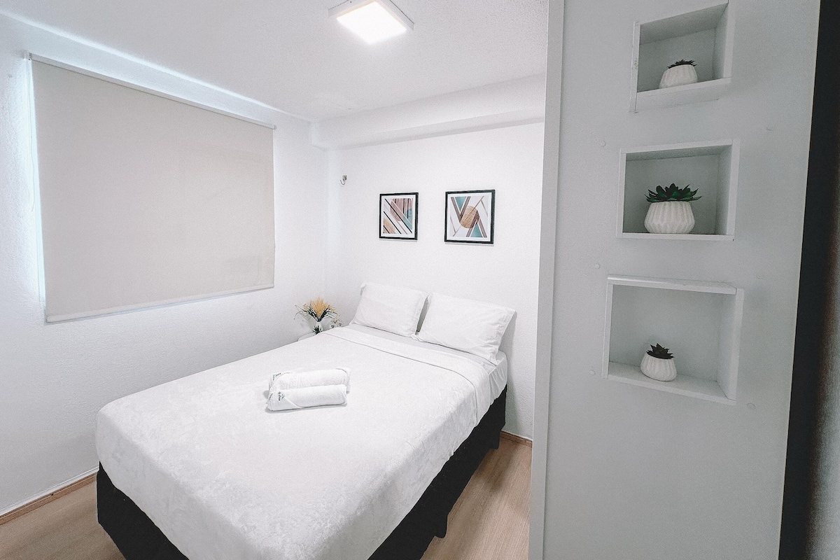 A bedroom with a bed, a window, a wall-mounted shelf with plants, and two framed pictures.