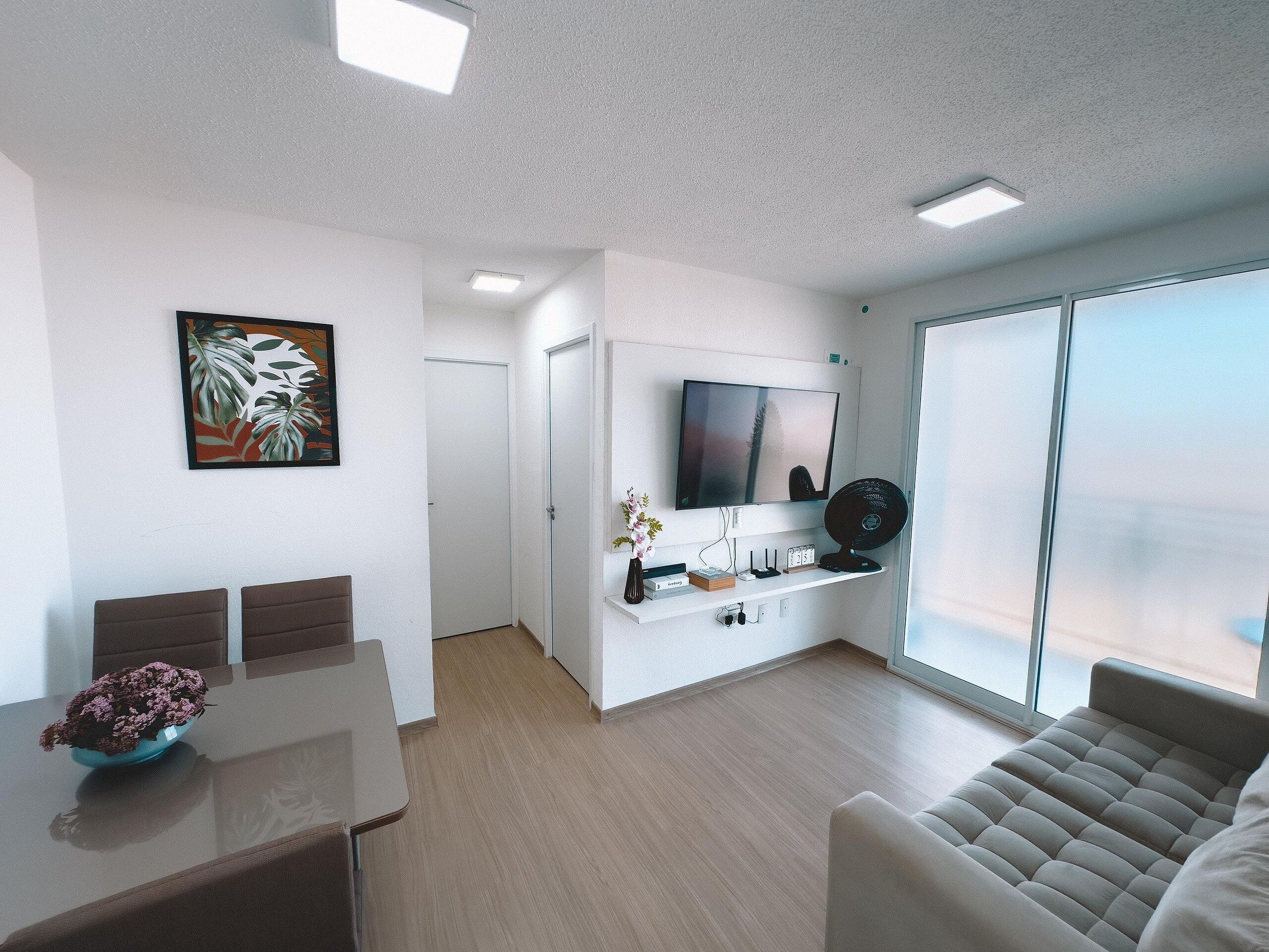 Una sala de estar moderna con un televisor de pantalla plana, un sofá, una mesa de comedor con un jarrón de flores y un ventanal grande.