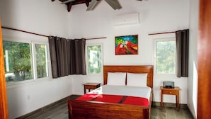 Un dormitorio con una cama de madera, dos mesitas de noche, un cuadro en la pared y ventanas grandes con cortinas.
