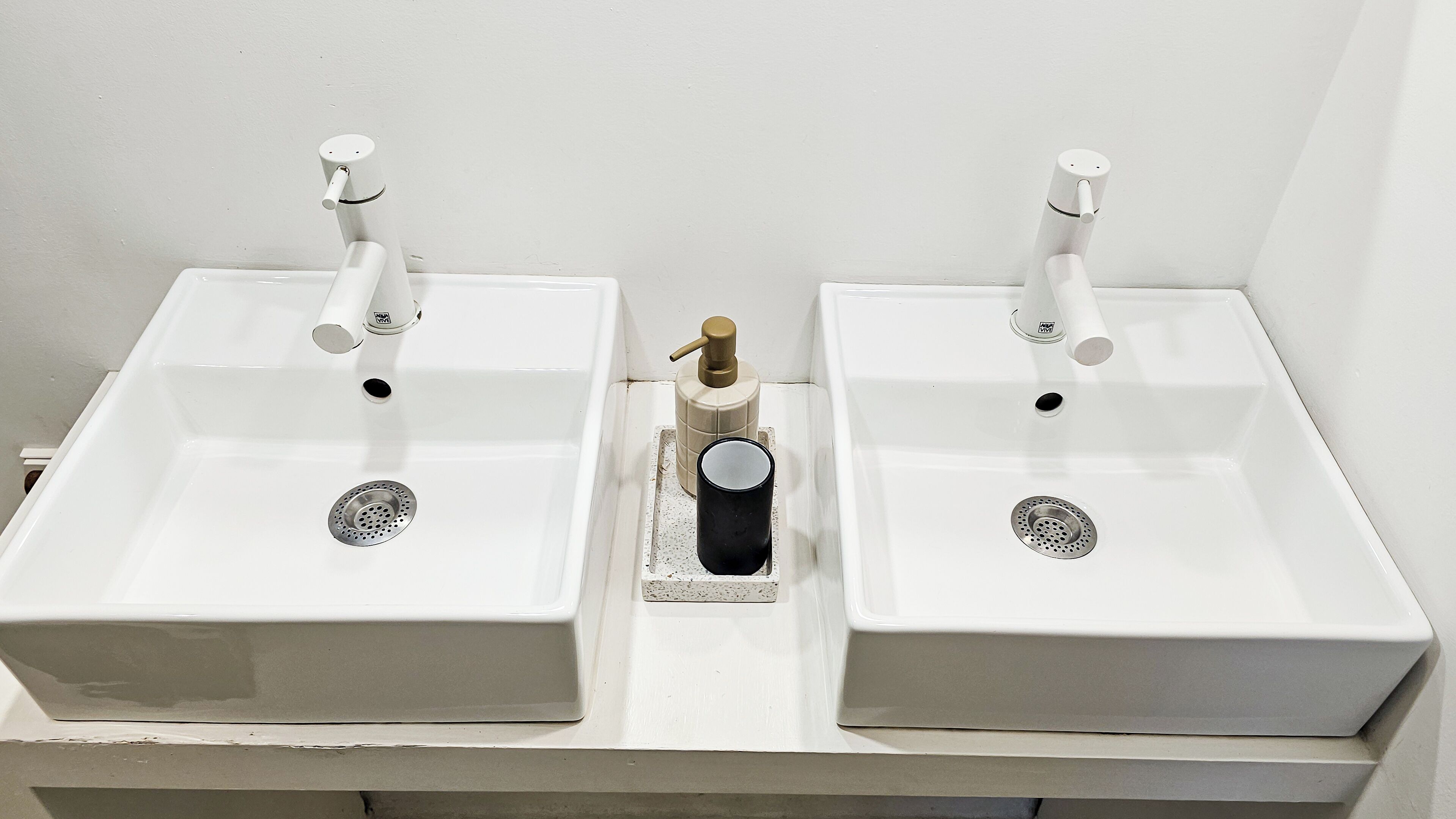 Twee witte wastafels met afzonderlijke kraanwerk en een zeepdispenser ertussen.