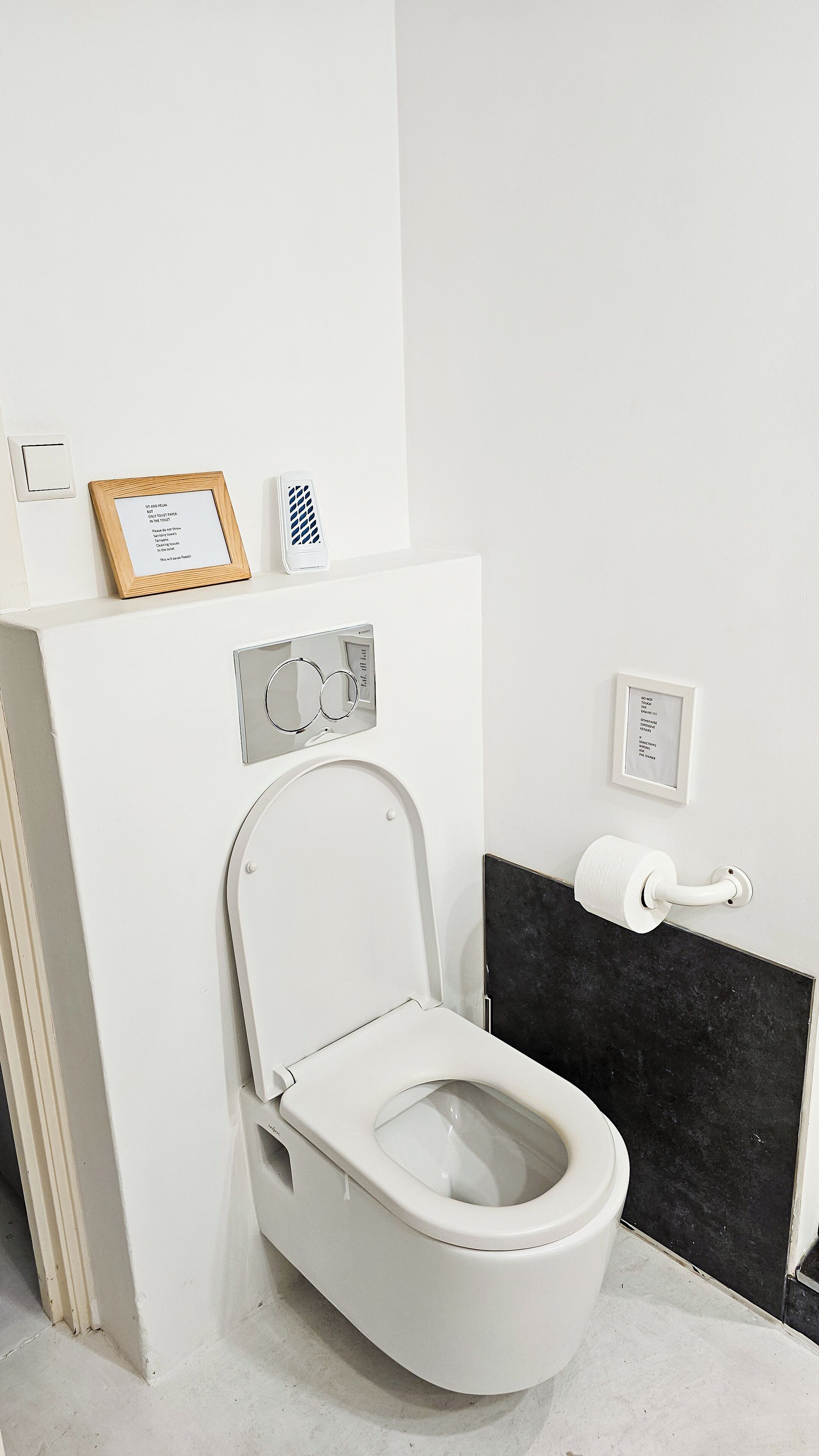 A modern bathroom with a white toilet, a black tiled wall, a framed sign, and a small white device on the wall.