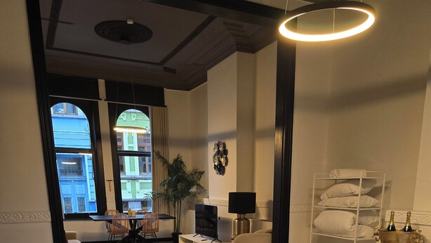 Una habitación de hotel moderna con una cama grande, una zona de estar con sofá y sillones, un televisor, una lámpara y vistas a la ciudad a través de amplias ventanas.