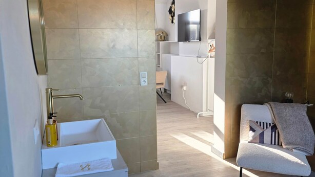 A modern bathroom with a sink, towel rack, and a mirror. There is a doorway leading to a living area with a sofa and a television.