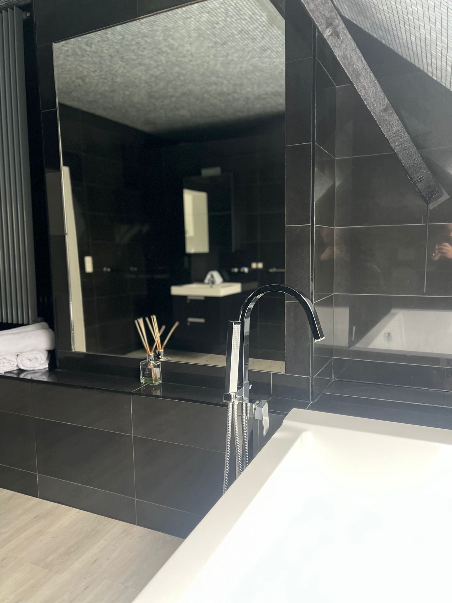 A modern bathroom with a large mirror, a black tiled wall, a white sink, and a bathtub.