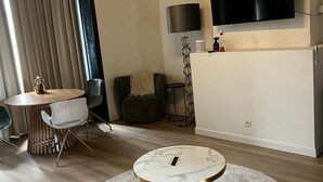 Una sala moderna con una mesa de centro redonda, un televisor de pantalla plana montado en la pared, una zona de comedor con una mesa redonda y sillas, y un ventanal con cortinas.
