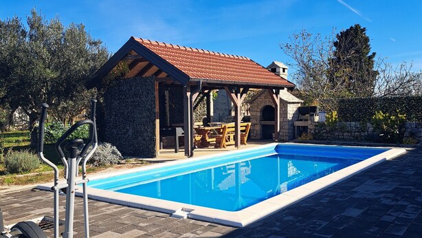 A swimming pool with a stone deck, a covered seating area, and a stone building with a tiled roof.