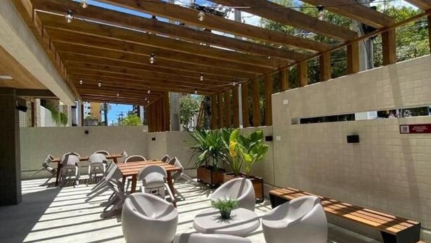 An outdoor seating area with wooden benches and white chairs, surrounded by a low wall and potted plants.