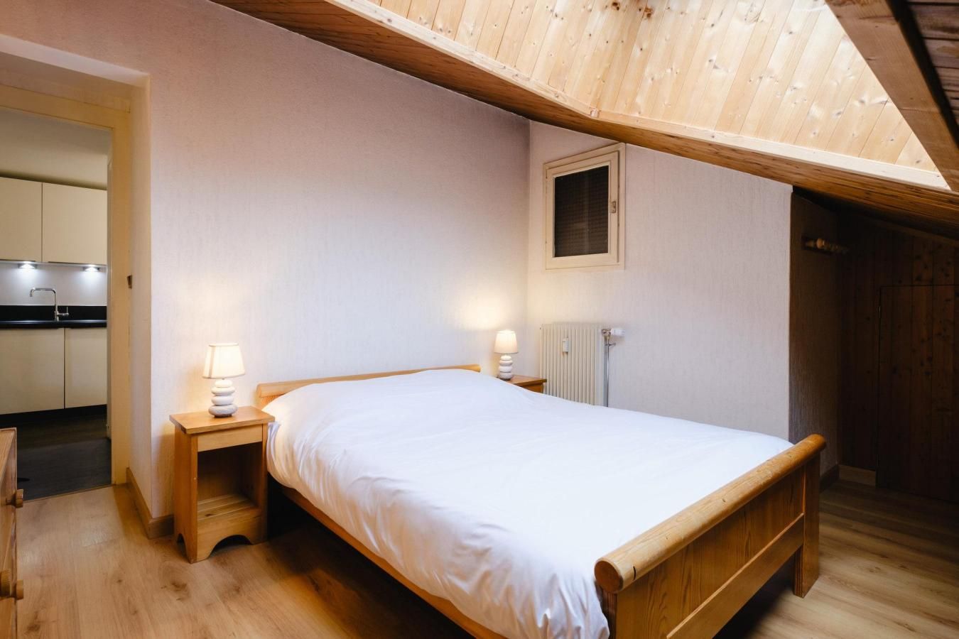 Un dormitorio con una cama de madera, dos mesitas de noche con lámparas, una ventana y una cocina visible a través de una puerta abierta.