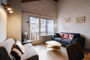 A cozy living room with a dark blue sofa, a wooden coffee table, and a white armchair. A large window offers a view of a snowy landscape. - 50m from ski slopes, 3rd floor, view village/mountain, balcony, tv, 76m², La Clusaz (La Clusaz)