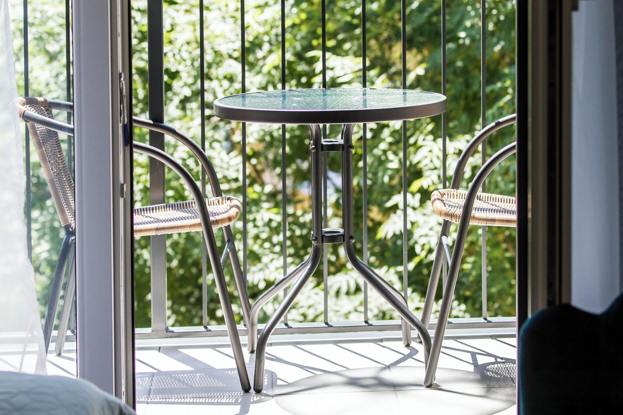 Een balkon met een ronde tafel en twee rieten stoelen, met uitzicht op een weelderig groen gebied.