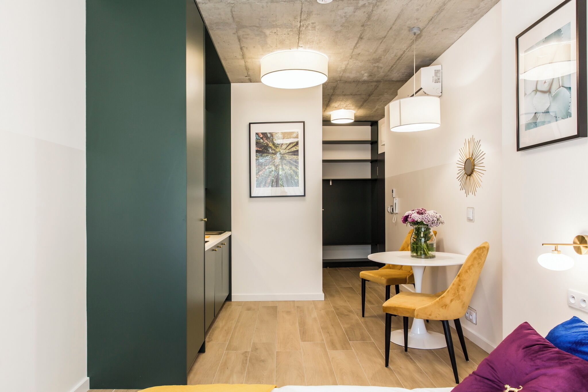 Une cuisine moderne avec un coin repas, comprenant une table ronde entourée de chaises jaunes, un mur blanc orné d’un tableau encadré et un plafond laissant apparaître le béton brut.