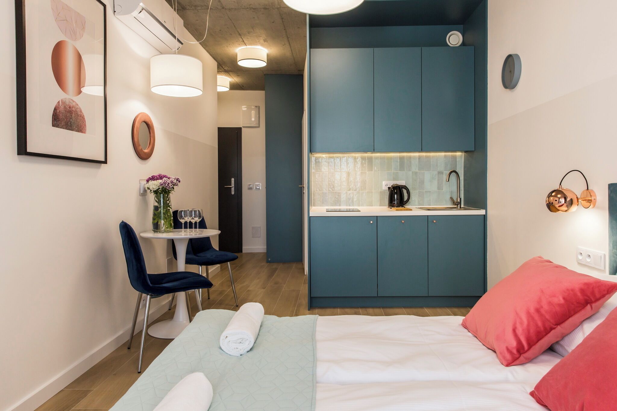 A compact hotel room with a bed, a small desk with chairs, a kitchenette, and a bathroom.