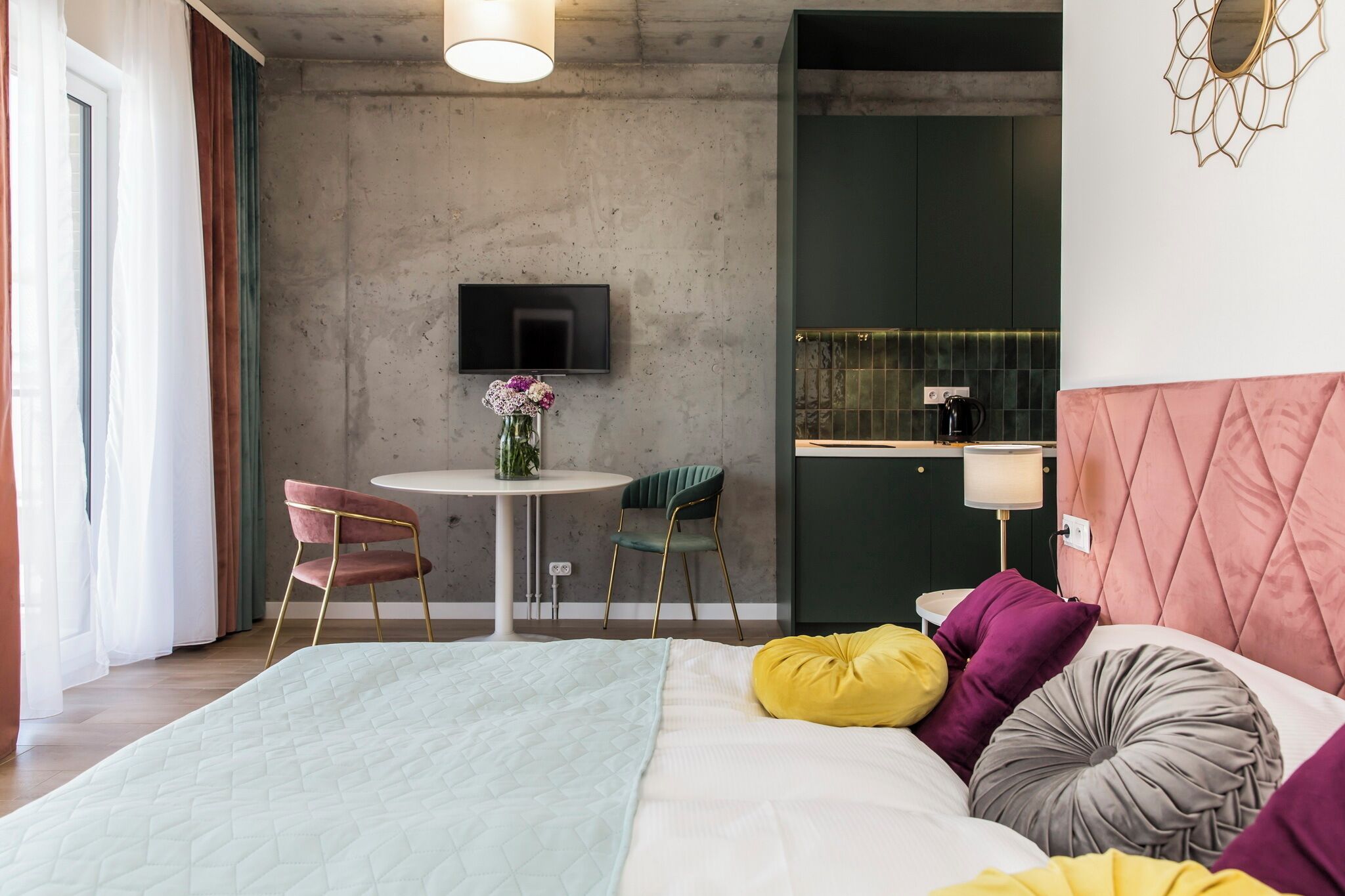 A modern bedroom with a bed, a bedside table, a TV, and a small dining area with chairs.