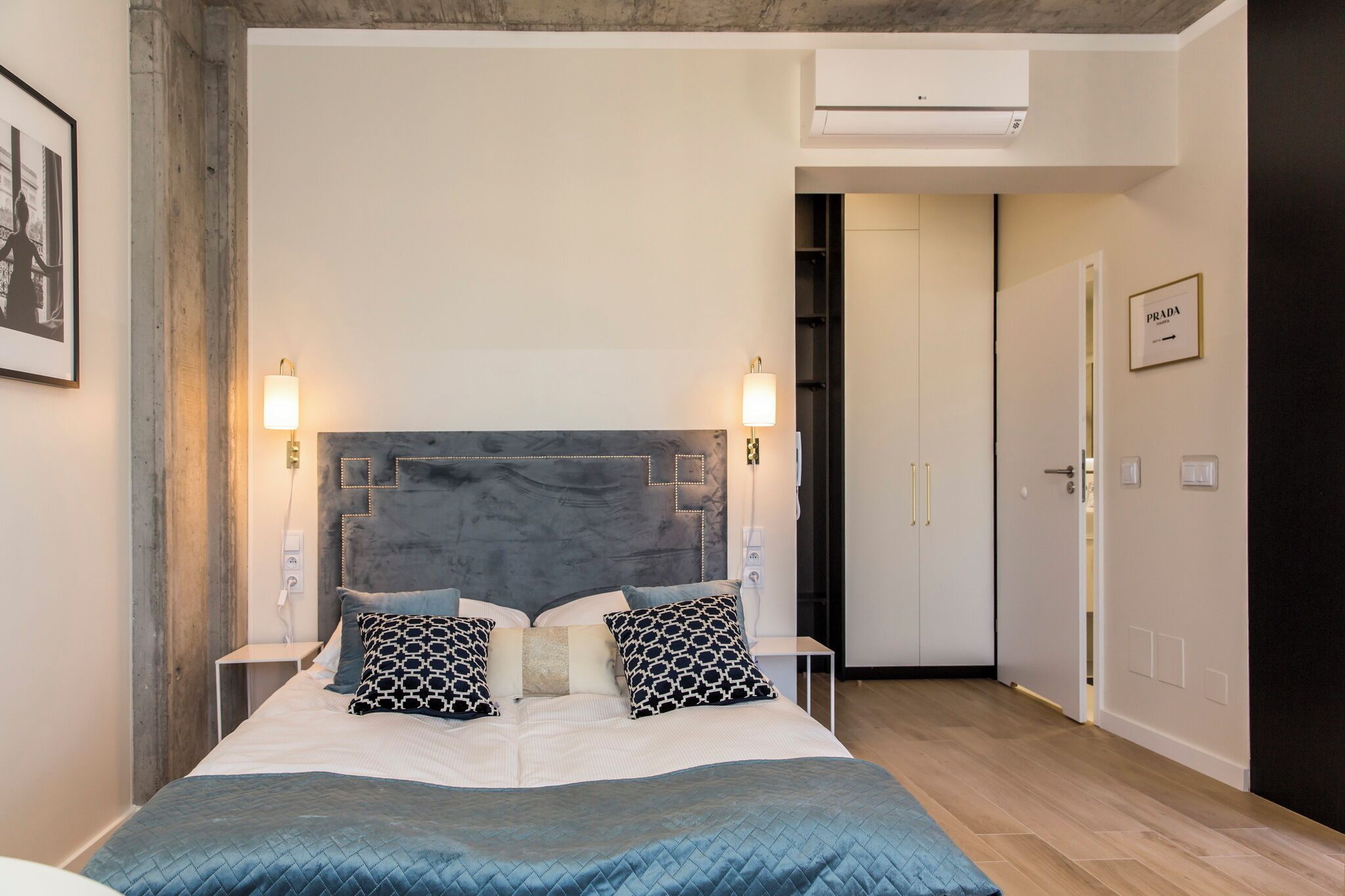 Quarto com cama, luminárias de cabeceira, guarda-roupa e porta com placa.