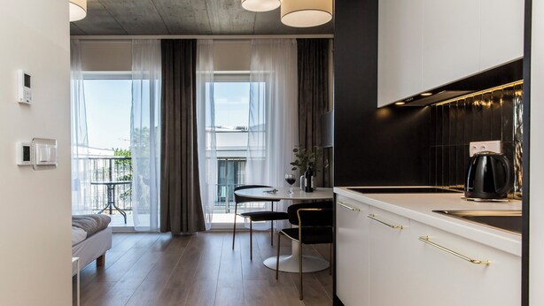 A modern kitchen with white cabinets and gold handles, a dining area with a round table and chairs, and a balcony with sheer curtains. - Piedmont House (Kraków)