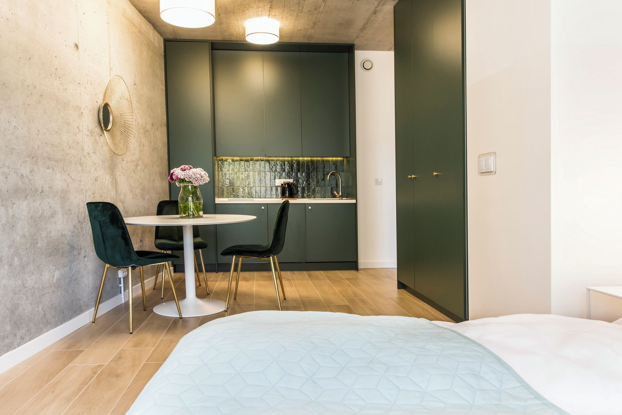 Una camera compatta con un letto, un tavolo rotondo da pranzo con due sedie, un angolo cottura con una finestra e uno specchio decorativo al muro.