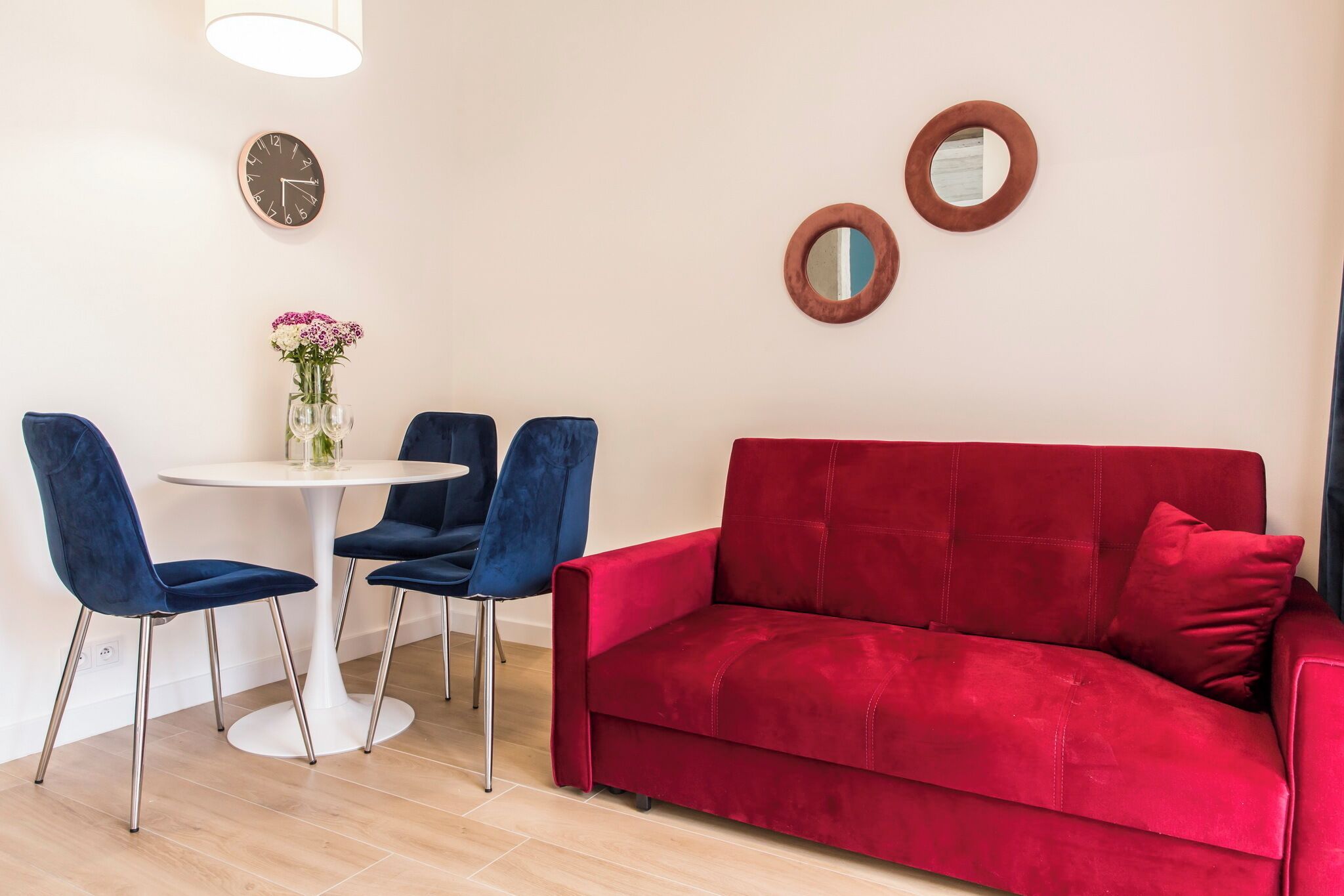 Ein modernes Wohnzimmer mit einem roten Sofa, blauen Sesseln, einem weißen Tisch und einer Uhr an der Wand.