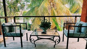 A wooden deck with two cushioned chairs and a glass-top table, surrounded by lush greenery and a thatched roof structure.
